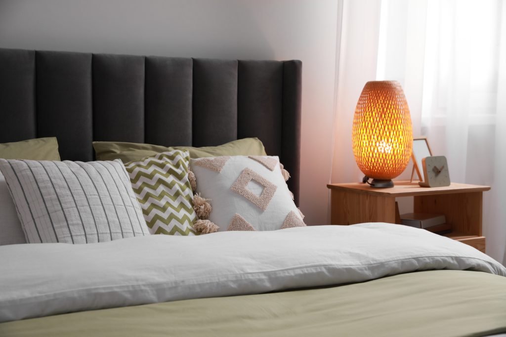 Comfortable bed with cushions, lamp and different decor on wooden bedside table in room. Stylish interior